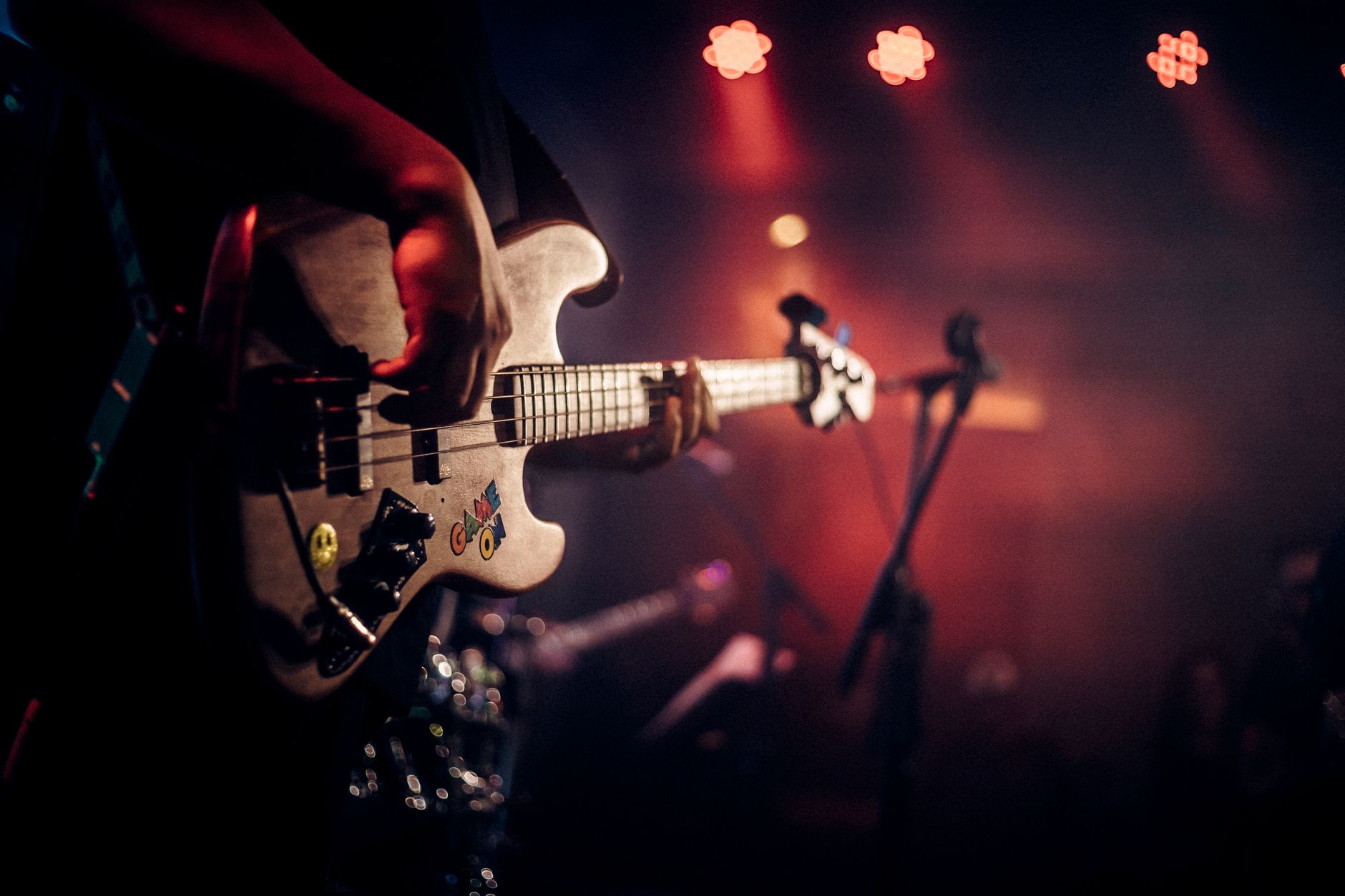 Main d’un musicien jouant d’une basse à autocollants sur une scène sombre, avec projecteurs rouges et pied de micro flous à l’arrière-plan.