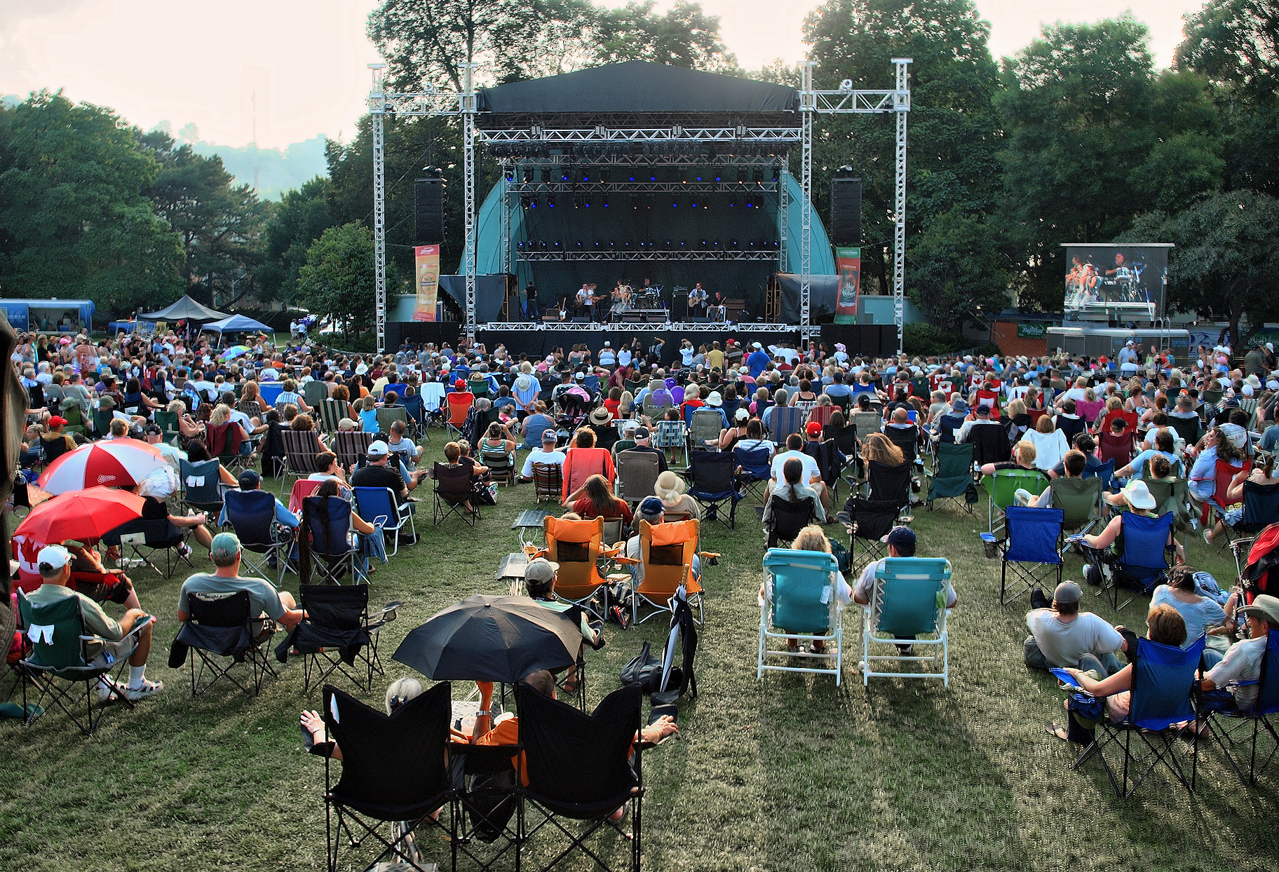 Public assis sur des chaises pliantes face à une scène de concert dressée sur la pelouse d’un parc arboré