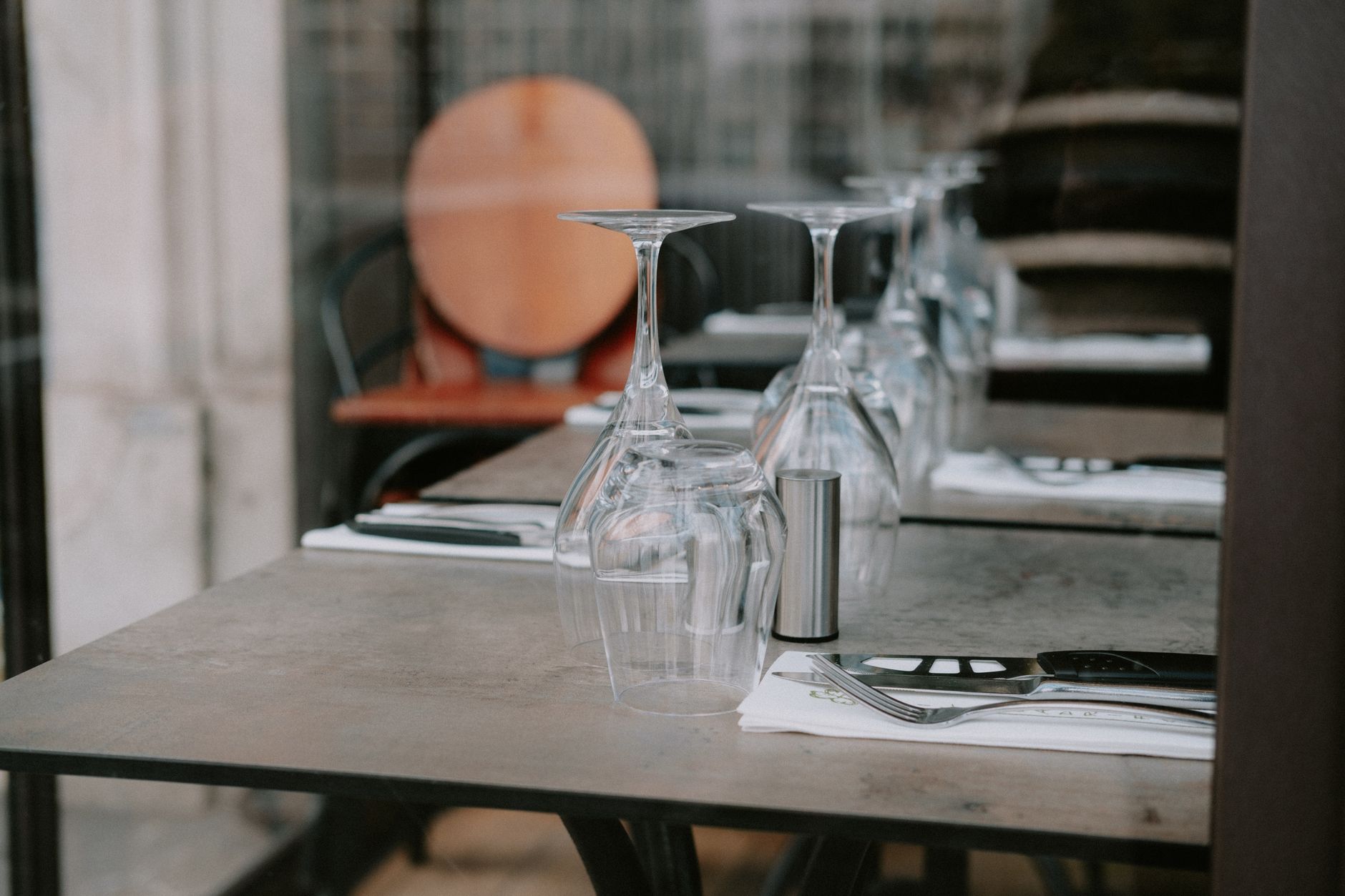 Tables de restaurant dressées avec verres à pied retournés, couverts sur serviettes blanches et chaise en bois floue en arrière-plan.