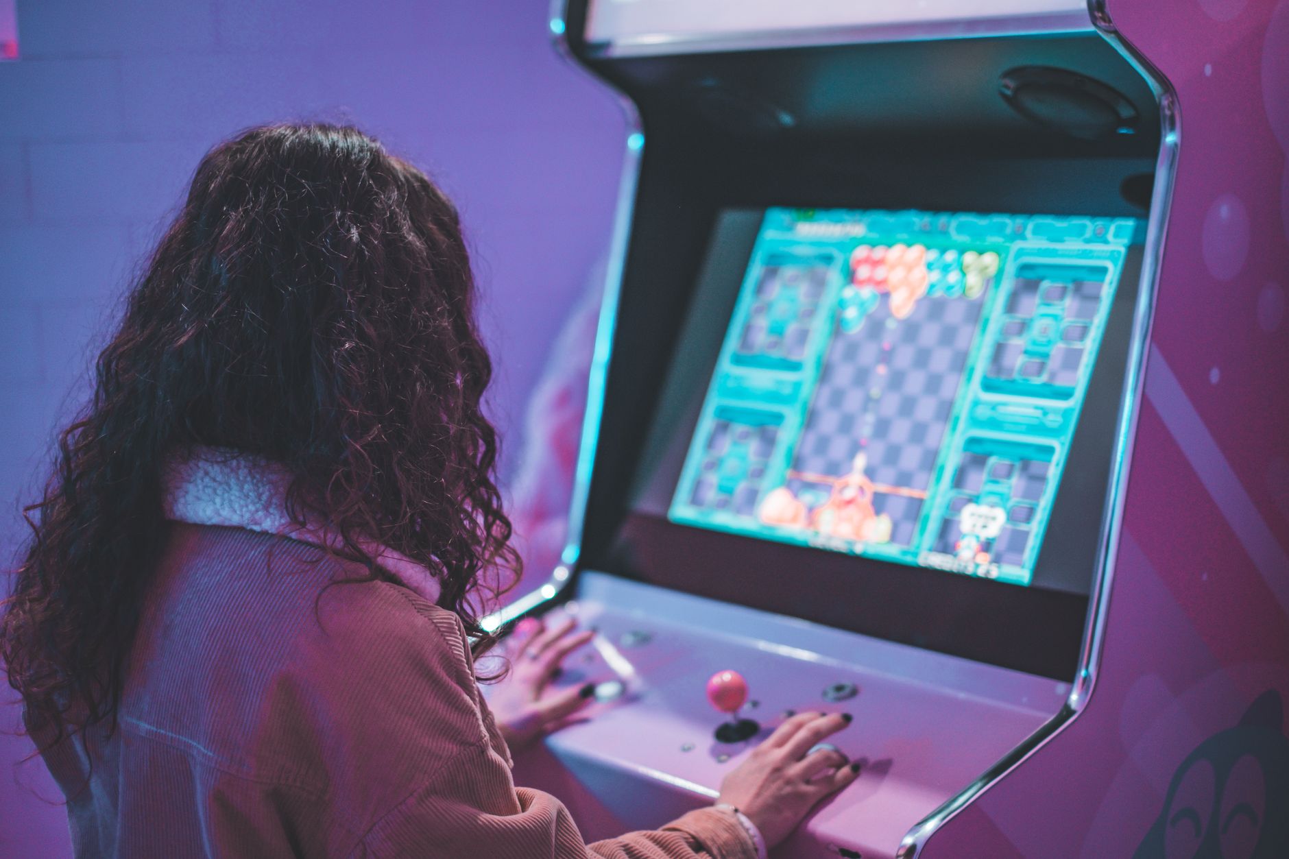 Une personne aux cheveux bouclés, vue de dos, joue sur une borne d’arcade dans une salle aux lumières violettes et roses.